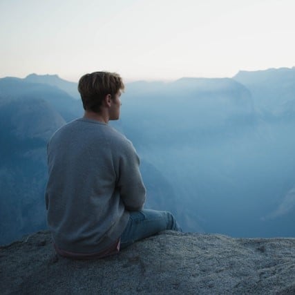 A man sitting on a cliff looking out at the mountains.
